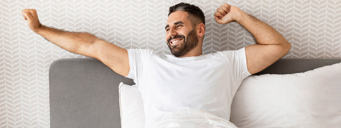 Man stretching and smiling in bed, wearing a white t-shirt.