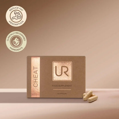 A box of UR Cheat Food Supplement capsules on a beige surface with a matching beige background. The box is brown with a metallic rose gold vertical stripe on the left and a metallic square logo on the front.