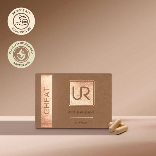 A box of UR Cheat Food Supplement capsules on a beige surface with a matching beige background. The box is brown with a metallic rose gold vertical stripe on the left and a metallic square logo on the front.