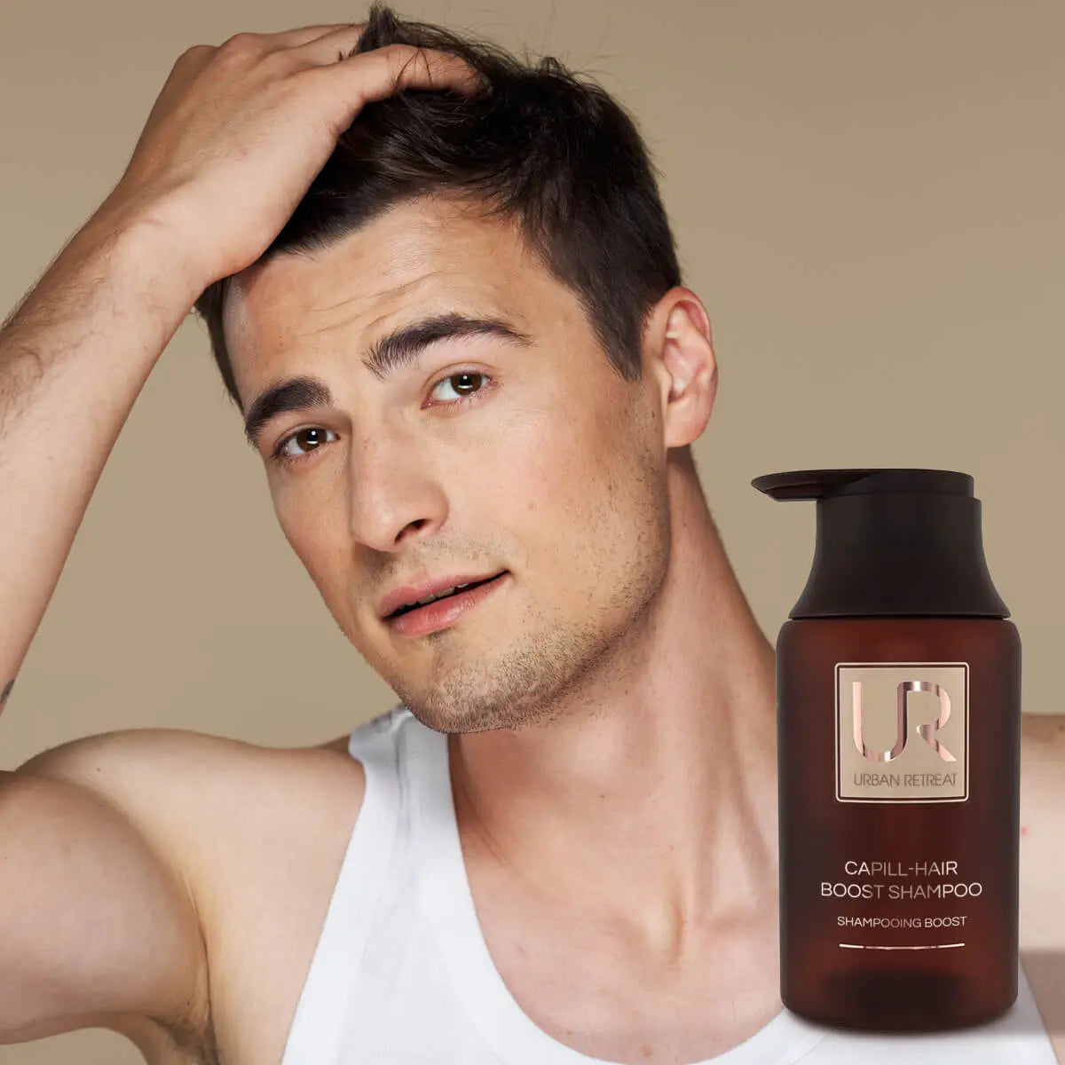 A man with short dark hair looks at the camera while touching his hair, wearing a white tank top. A brown pump bottle labeled UR Captive-Hair Boost Shampoo is shown in the lower right corner against a plain beige background.