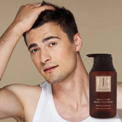 A man with short dark hair looks at the camera while touching his hair, wearing a white tank top. A brown pump bottle labeled UR Captive-Hair Boost Shampoo is shown in the lower right corner against a plain beige background.