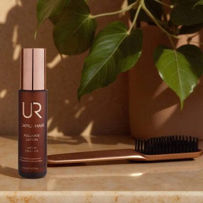 UR Capili-Hair Follviate Lotion bottle with a metallic rose gold cap, placed on a marble surface next to a matching rose gold hairbrush, with green leaves and a brown pot in the background.