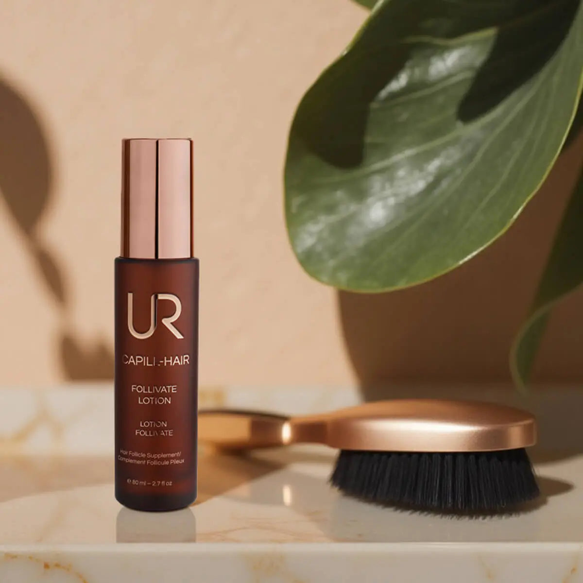 UR Capili-Hair Follviate Lotion bottle in dark brown with a metallic rose gold cap, standing on a marble surface next to a rose gold and black hairbrush, with a green plant leaf in the background.